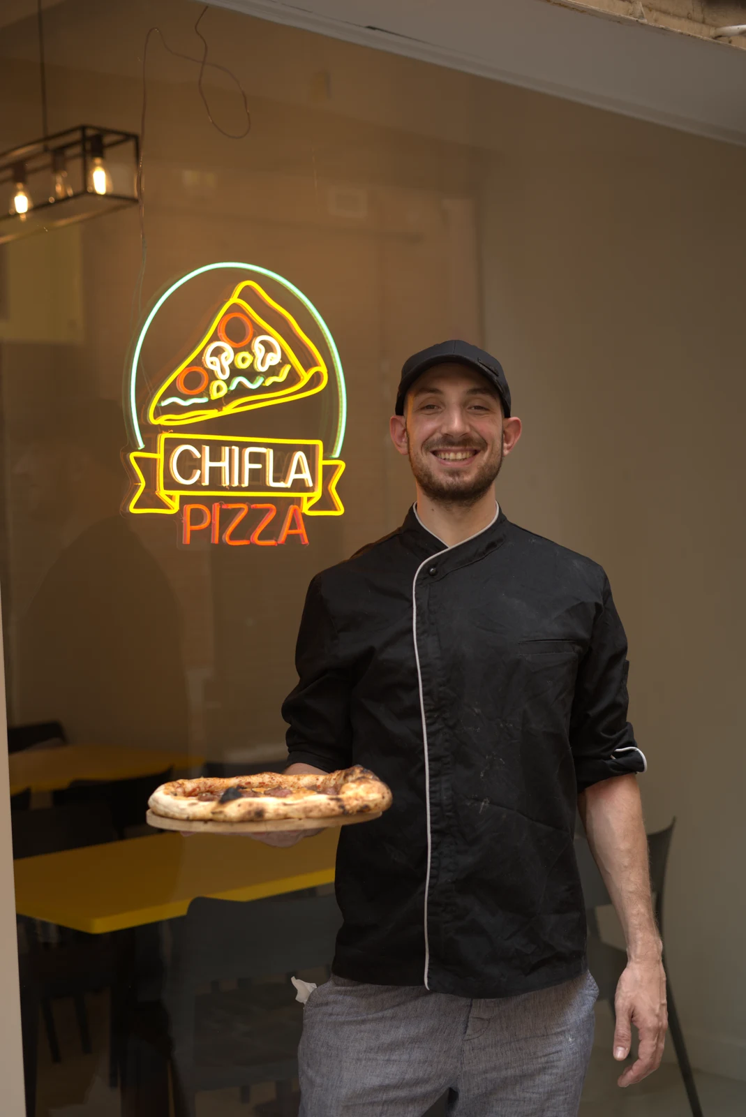 Pizzaiolo de Chifla sosteniendo una pizza junto al neón del local